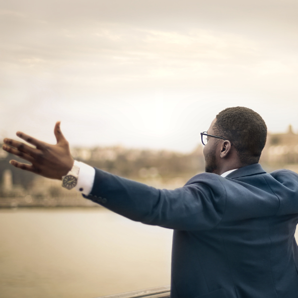 An gentleman with dark brown skin, short hair, and glasses faces a river and landscape. He is wearing a suit and has his arms outreached in a feeling of joy.